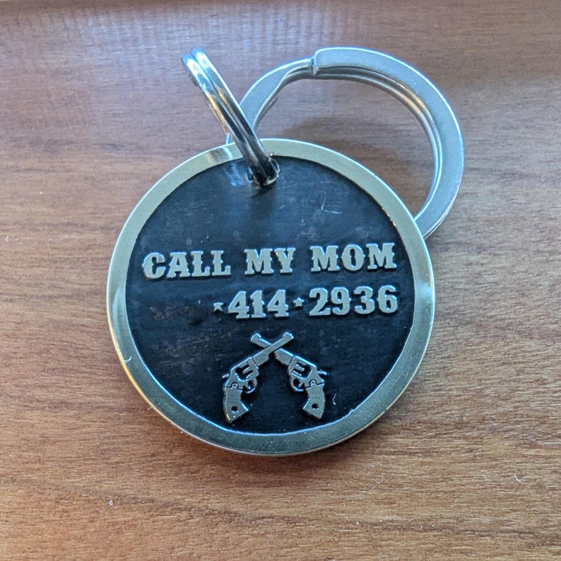 May include: A circular silver-toned metal pet ID tag with a black recessed center. The text "CALL MY MOM" is engraved above the number "414-2936" and a graphic of two crossed revolvers. A silver split ring is attached to the top of the tag.