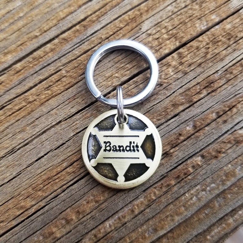 May include: A round brass-toned metal pet ID tag featuring a six-pointed star design with a dark recessed background. The word Bandit is engraved in black lettering across the center of the star. The tag is attached to a silver-toned metal split ring.