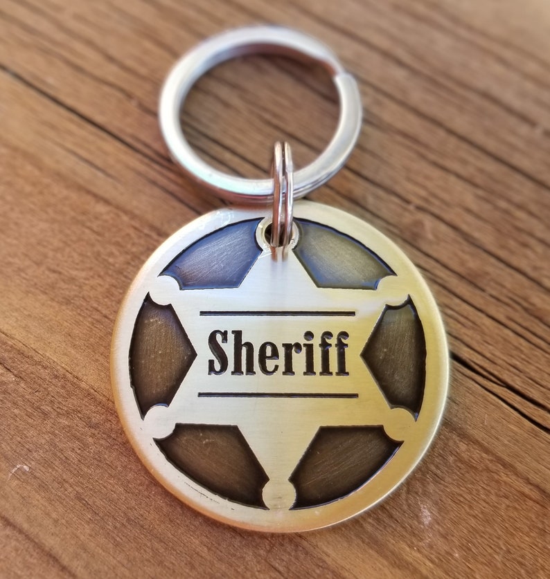 May include: A circular brass-toned metal keychain tag featuring a six-pointed star cutout design. The word Sheriff is engraved in black lettering across the center of the star. The tag is attached to a silver-colored metal split ring for keys.