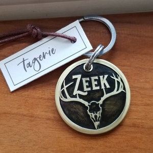 May include: A round brass dog tag with a black background and a gold rim. The tag features a stylized deer skull with antlers and the name "ZEek" in gold lettering. The tag is attached to a silver key ring.