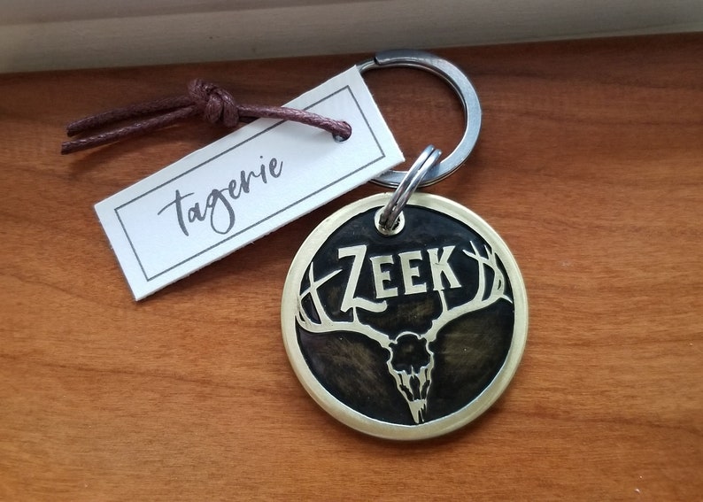 May include: A round brass dog tag with a black background and a gold rim. The tag features a stylized deer skull with antlers and the name "ZEek" in gold lettering. The tag is attached to a silver key ring.