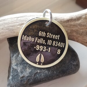 May include: A round, brass key chain tag with a dark brown background. The tag has the text "6th Street Idaho Falls, ID 83401 -993-1 8" engraved on it. The tag also has a gold-colored elk hoof print.