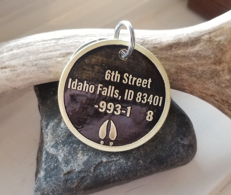 May include: A round, brass key chain tag with a dark brown background. The tag has the text "6th Street Idaho Falls, ID 83401 -993-1 8" engraved on it. The tag also has a gold-colored elk hoof print.