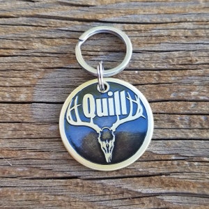 May include: A gold-colored metal keychain with a black background featuring a deer skull with antlers and the word "Quill" in white lettering.
