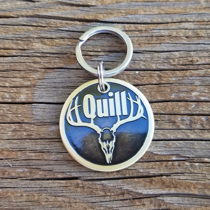 May include: A gold-colored metal keychain with a black background featuring a deer skull with antlers and the word "Quill" in white lettering.