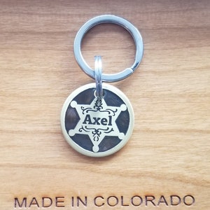 May include: A brass dog tag with a five-pointed star and the name "Axel" engraved in a decorative font. The tag is attached to a silver key ring. The tag is on a wooden surface with the words "MADE IN COLORADO" engraved below it.