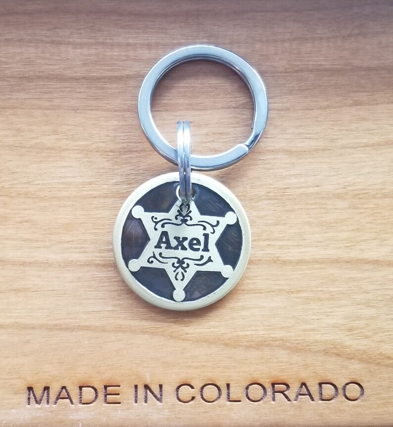 May include: A brass dog tag with a five-pointed star and the name "Axel" engraved in a decorative font. The tag is attached to a silver key ring. The tag is on a wooden surface with the words "MADE IN COLORADO" engraved below it.