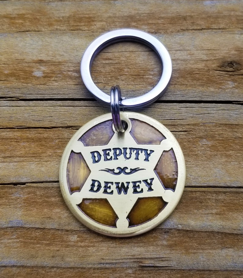 May include: A gold-coloured metal keychain featuring a sheriff's star design. The star has a brown wood-grain pattern and the words "DEPUTY DEWEY" are engraved in black.