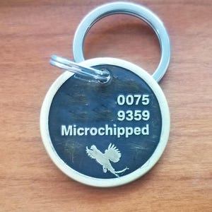 May include: A round, silver-colored pet tag with a black background. The tag has the numbers 0075 and 9359, the word "Microchipped", and a silhouette of a bird.