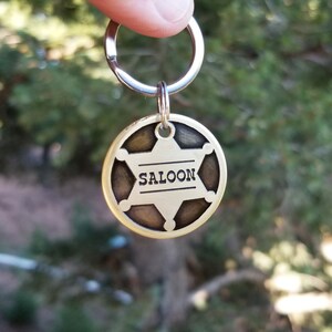 May include: A circular brass-toned metal keychain featuring a six-pointed star design with a dark, recessed background. The word SALOON is engraved in black capital letters across the center of the star, which is attached to a silver-toned ring.