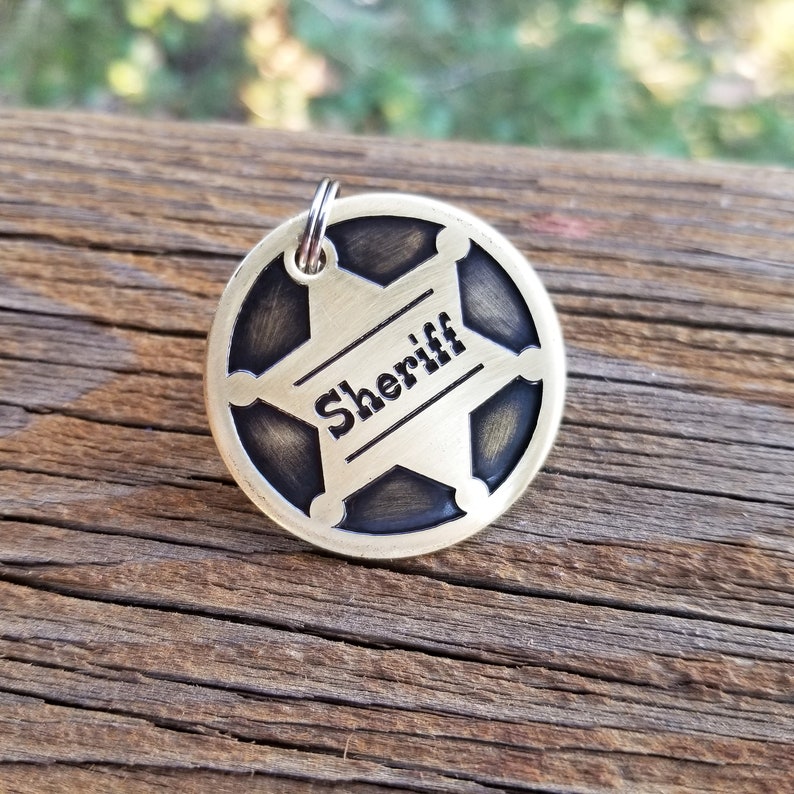 May include: A circular brass-toned metal tag featuring a recessed five-pointed star design with the word Sheriff engraved in the center. The tag has a dark, antiqued finish within the star and includes a small silver-toned metal jump ring at the top.