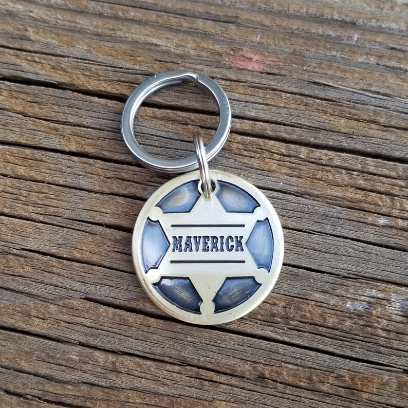 May include: A circular metal pet tag featuring a six-pointed star design with a dark, recessed finish. The name MAVERICK is engraved in black capital letters across the center of the star. A silver-toned metal split ring is attached to the top.