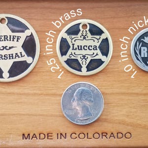 May include: Three circular metal pet tags with holes for attachment. The 1.5 inch brass tag features a sheriff star and the text SHERIFF MARSHAL. The 1.25 inch brass tag has a star and the name Lucca. The 1.0 inch nickel tag shows birds and the name Remi.