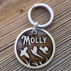 May include: A round, brass dog tag with a brown woodgrain finish. The tag features the name "MOLLY" in a stylized font and three pheasant silhouettes. The tag is attached to a silver split ring.