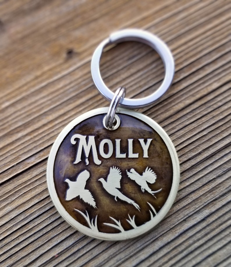 May include: A round, brass dog tag with a brown woodgrain finish. The tag features the name "MOLLY" in a stylized font and three pheasant silhouettes. The tag is attached to a silver split ring.