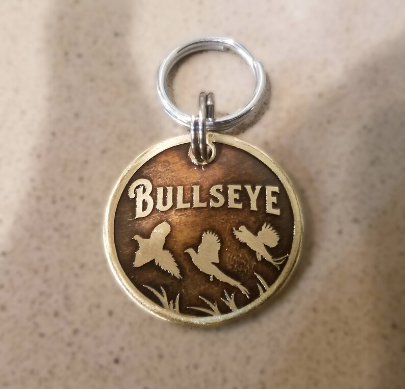 May include: Brass dog tag with a brown background and the word "BULLSEYE" in gold lettering. Three pheasant silhouettes are flying in front of the text.