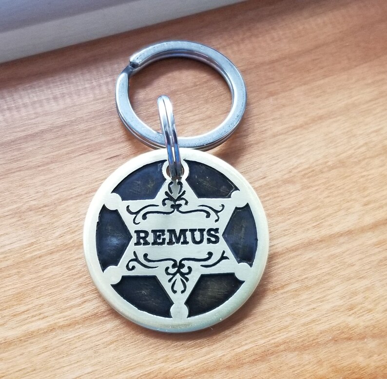 May include: A brass dog tag with a five-pointed star and the name "REMUS" engraved in the center. The tag is attached to a silver key ring.