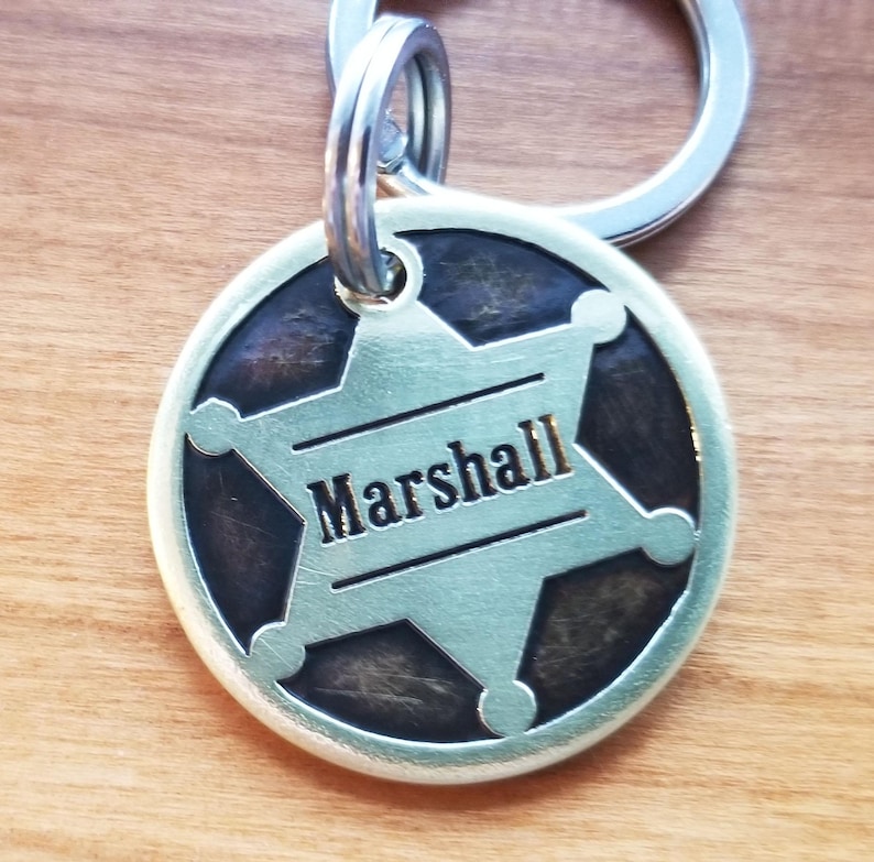 May include: A circular silver-toned metal pet tag featuring a six-pointed sheriff star emblem centered on a dark, textured background. The name Marshall is engraved in a bold, black sans-serif font across the middle of the star. A metal ring is attached.