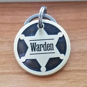 May include: A circular metal keychain tag featuring a six-pointed star emblem with a dark background. The word Warden is engraved in black capital letters across the center of the star. The tag includes a silver-toned metal split ring at the top.