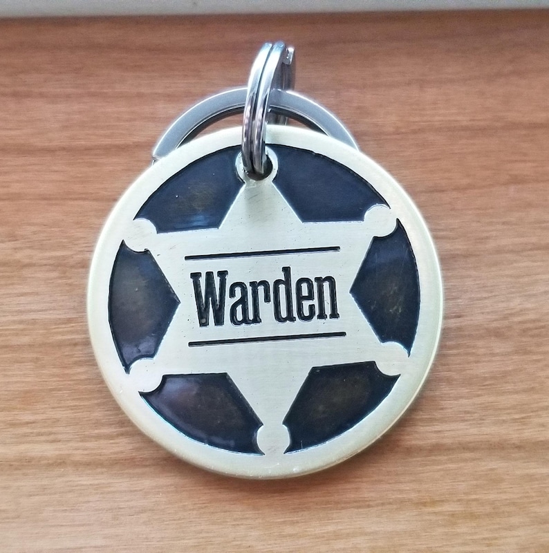 May include: A circular metal keychain tag featuring a six-pointed star emblem with a dark background. The word Warden is engraved in black capital letters across the center of the star. The tag includes a silver-toned metal split ring at the top.