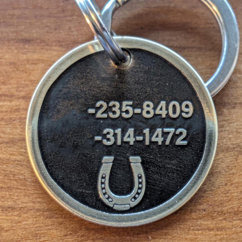 May include: A round metal pet identification tag with a dark, textured center and a lighter metallic rim. The tag features the engraved numbers -235-8409 and -314-1472, along with a horseshoe icon at the bottom. A metal split ring is attached to the top.