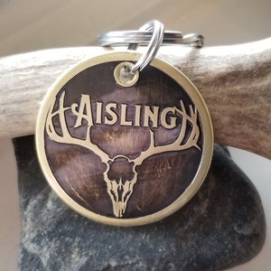 May include: Brass dog tag with a brown background and a gold deer skull with antlers. The name "Aisling" is engraved in gold lettering.