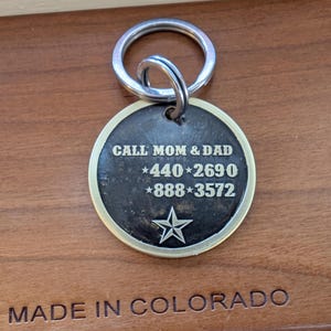 May include: A round, dark-toned metal pet identification tag with a silver-colored rim and a split ring. The tag features the engraved text CALL MOM & DAD, the phone numbers 440-2690 and 888-3572 separated by stars, and a single star at the bottom.