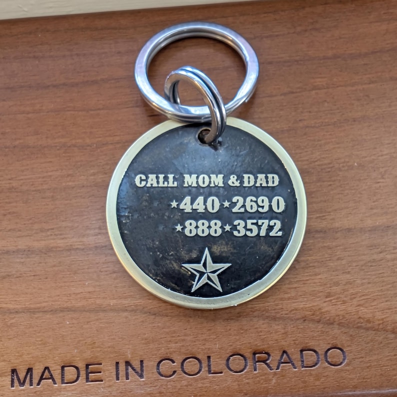 May include: A round, dark-toned metal pet identification tag with a silver-colored rim and a split ring. The tag features the engraved text CALL MOM & DAD, the phone numbers 440-2690 and 888-3572 separated by stars, and a single star at the bottom.