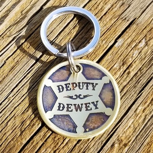 May include: A brass keychain with a silver key ring. The keychain features a round, five-pointed star-shaped tag with a blue and brown background. The tag says "DEPUTY DEWEY" in a stylized font.