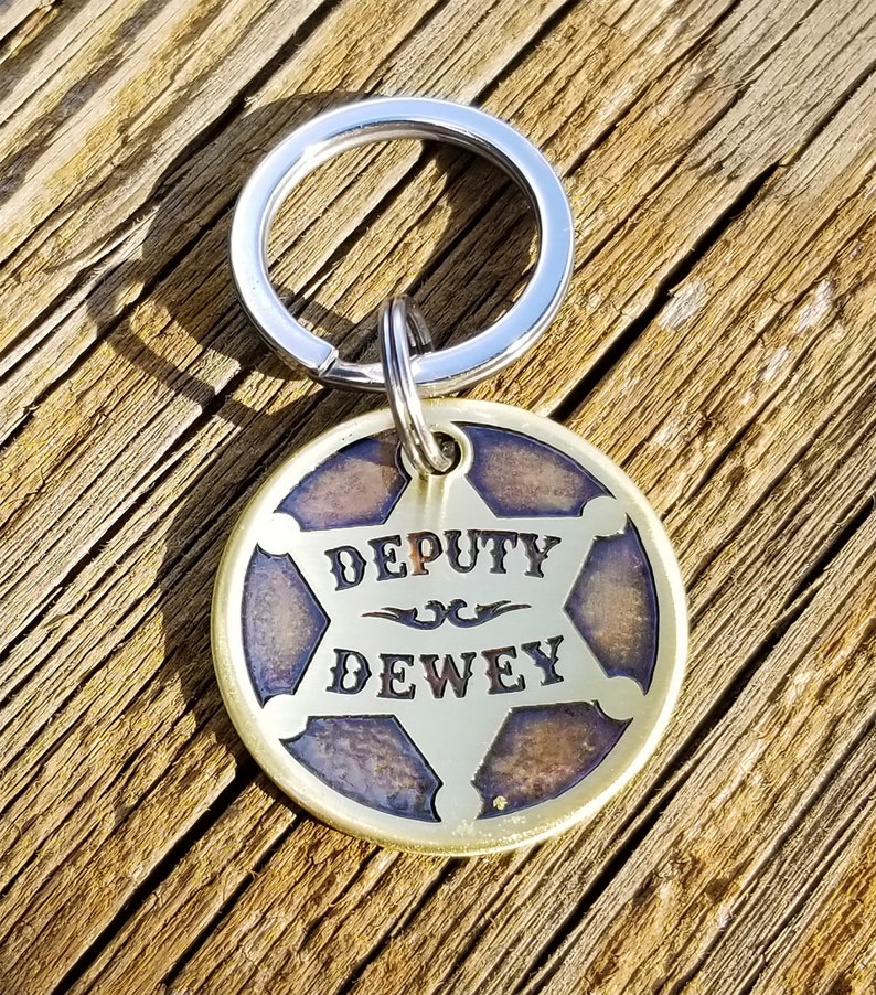 May include: A brass keychain with a silver key ring. The keychain features a round, five-pointed star-shaped tag with a blue and brown background. The tag says "DEPUTY DEWEY" in a stylized font.