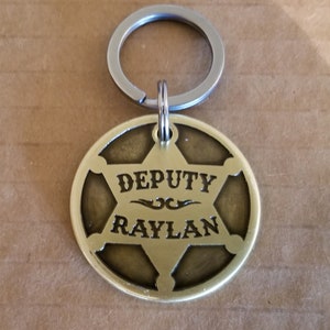 May include: A brass keychain featuring a sheriff's star design. The star is inscribed with the words "DEPUTY RAYLAN".