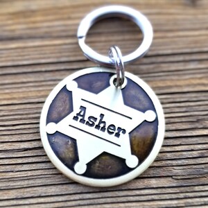 May include: A circular metal pet ID tag featuring a silver-toned sheriff star emblem set against a dark, mottled brown background. The name Asher is engraved in black lettering across the center of the star, and a silver split ring is attached at the top.