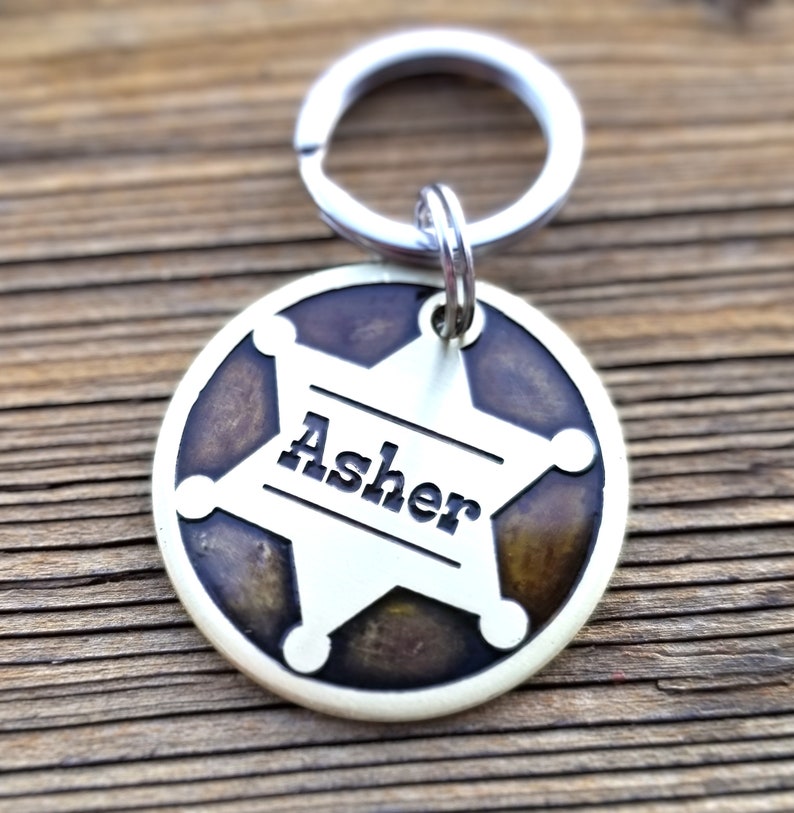 May include: A circular metal pet ID tag featuring a silver-toned sheriff star emblem set against a dark, mottled brown background. The name Asher is engraved in black lettering across the center of the star, and a silver split ring is attached at the top.