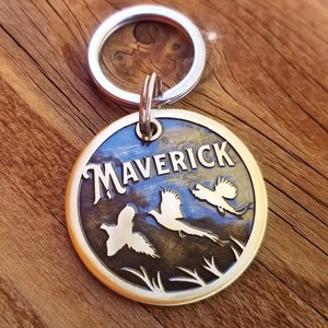 May include: Brass dog tag with a silver key ring. The tag is engraved with the name "Maverick" and three stylized flying birds.