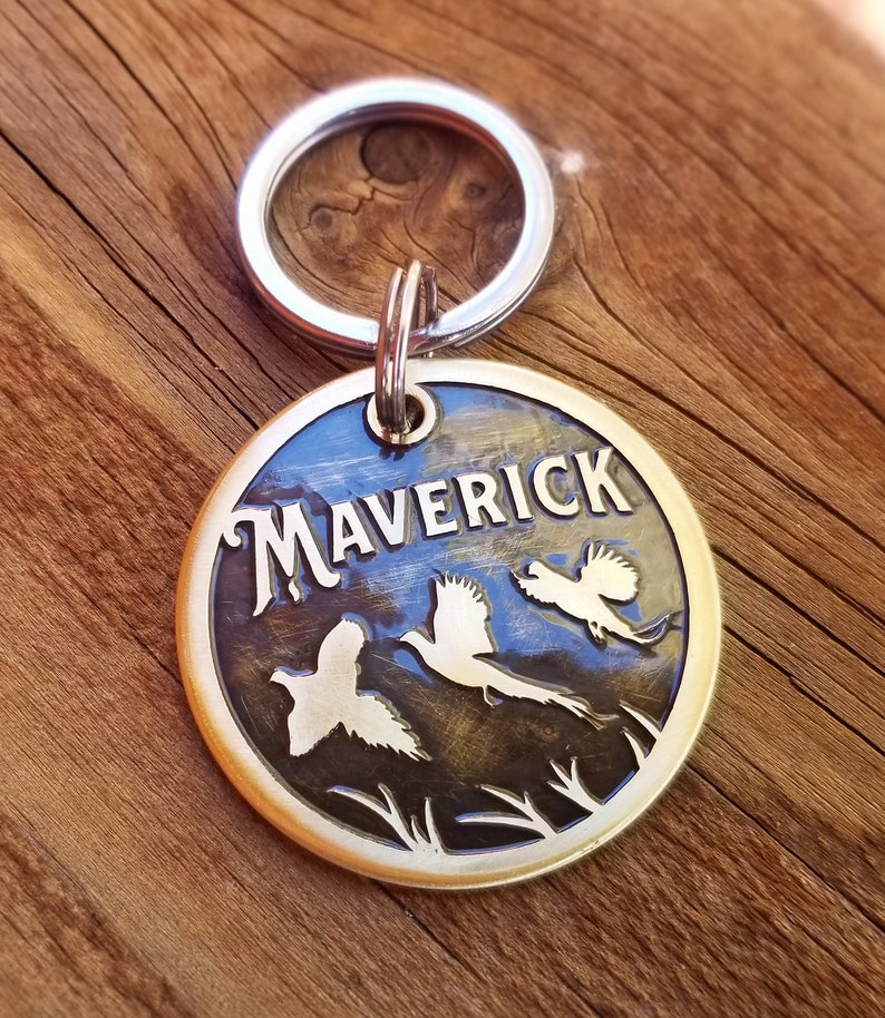 May include: Brass dog tag with a silver key ring. The tag is engraved with the name "Maverick" and three stylized flying birds.