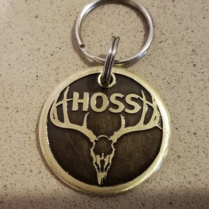 May include: A gold-colored metal dog tag with a black background. The tag features a stylized deer skull with antlers and the name "HOSS" in gold lettering.