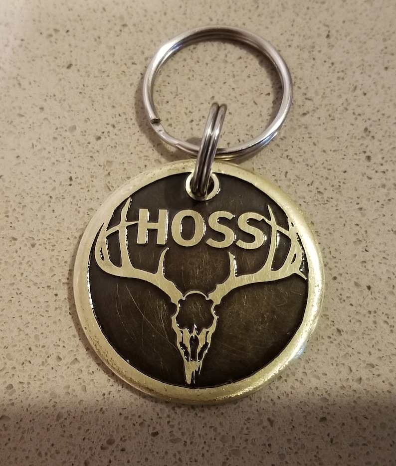 May include: A gold-colored metal dog tag with a black background. The tag features a stylized deer skull with antlers and the name "HOSS" in gold lettering.