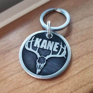 May include: A silver metal dog tag with a black background and a deer skull with antlers. The name "KANE" is engraved in white lettering on the tag.
