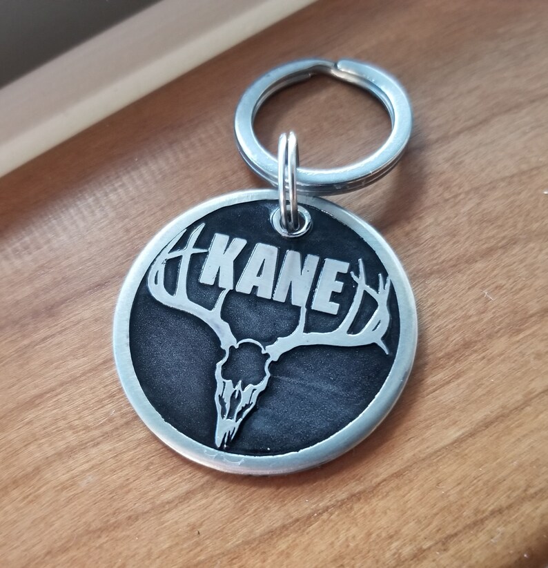 May include: A silver metal dog tag with a black background and a deer skull with antlers. The name "KANE" is engraved in white lettering on the tag.