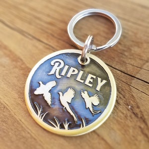 May include: Brass dog tag with a round shape and a silver key ring. The tag has a dark blue background with a gold inscription that reads "RIPLEY". Three white silhouettes of birds are etched into the tag.