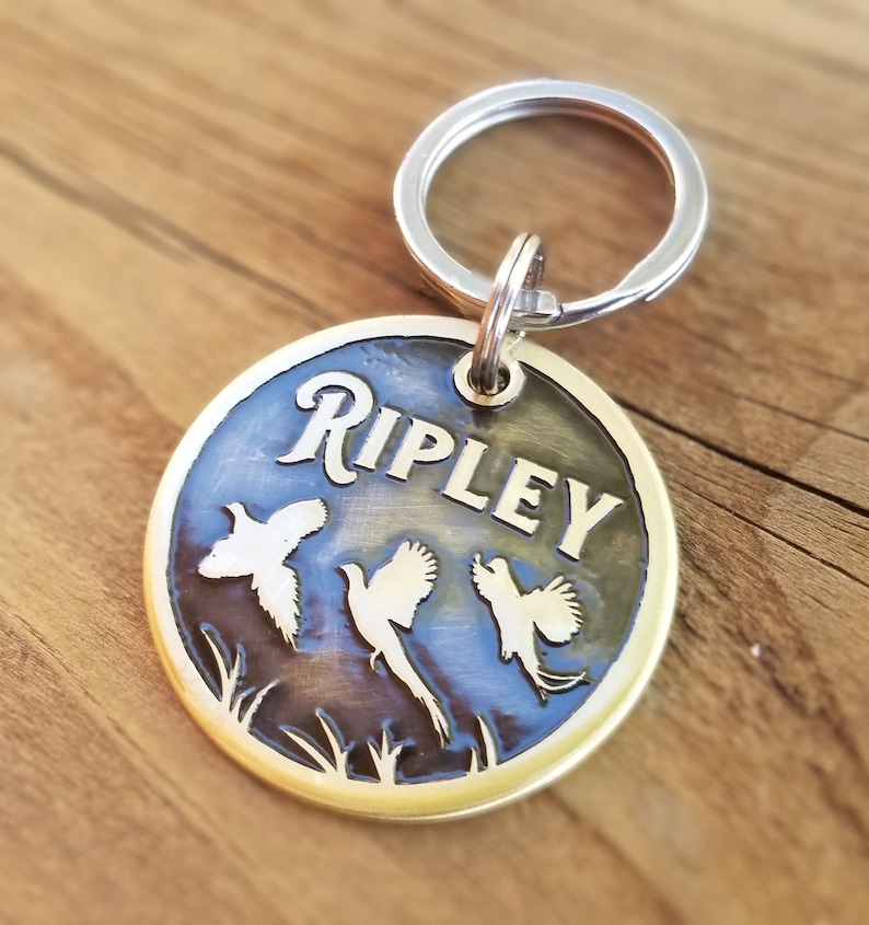 May include: Brass dog tag with a round shape and a silver key ring. The tag has a dark blue background with a gold inscription that reads "RIPLEY". Three white silhouettes of birds are etched into the tag.