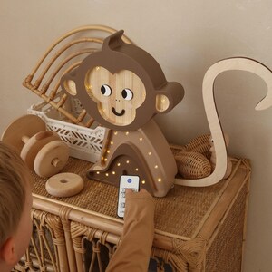 May include: A wooden monkey night light with a brown and beige color scheme. The light has a remote control and is lit up with warm white LED lights. The monkey is sitting on a wicker table.