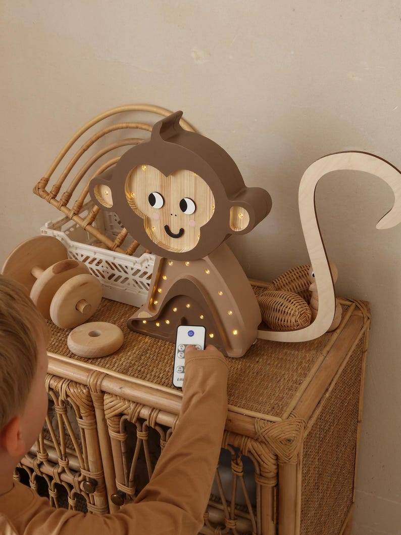 May include: A wooden monkey night light with a brown and beige color scheme. The light has a remote control and is lit up with warm white LED lights. The monkey is sitting on a wicker table.