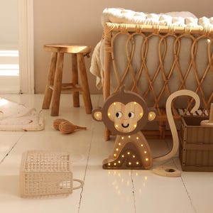 May include: A wooden monkey night light with a brown body and a light brown face. The monkey has a long tail and is sitting on a white wooden floor. The light is on and the monkey is glowing.