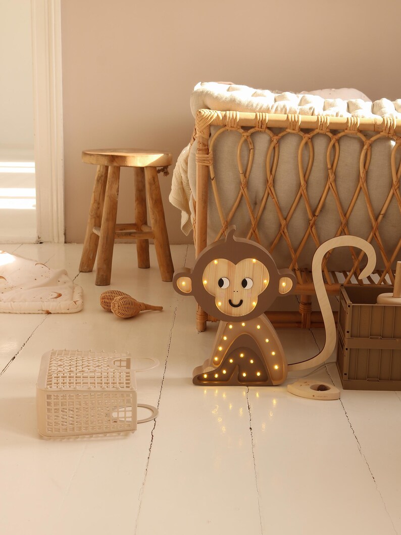 May include: A wooden monkey night light with a brown body and a light brown face. The monkey has a long tail and is sitting on a white wooden floor. The light is on and the monkey is glowing.