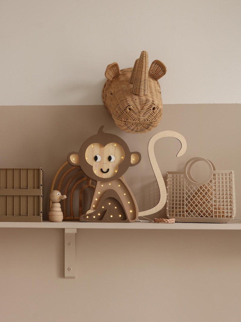 May include: A wooden monkey night light with a brown and tan finish and a smiling face. The monkey is sitting on a shelf with a woven rhino head, a wooden rainbow, and a woven basket.