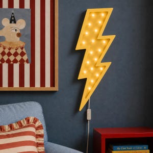 Könnte beinhalten: Eine gelbe Wandleuchte in Blitzform mit mehreren integrierten Glühbirnen. Die Lampe verfügt über ein weißes Stromkabel mit einem Schalter, das nach unten hängt. Die Blitzform ist beleuchtet und strahlt ein warmes Licht aus.