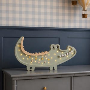 Op de afbeelding: Een lichtgroen houten nachtlampje in de vorm van een alligator met kleine, warme lampjes. De alligator heeft een houten kaak en tanden en staat op een grijze ladekast. Twee luchtballondecoraties zijn op de achtergrond.