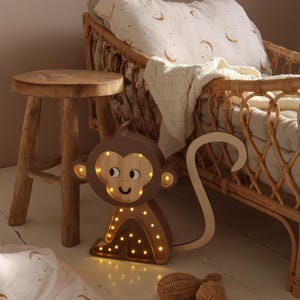 May include: A wooden monkey night light with a brown finish and a smiling face. The light has a long tail and is illuminated with warm white LED lights. The night light is sitting on a white wooden floor.
