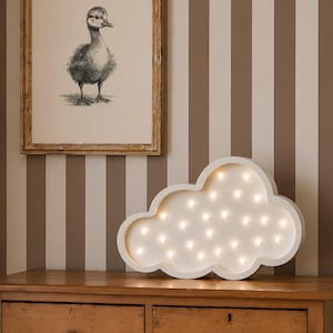 May include: A white cloud-shaped decorative light with multiple small bulbs, sitting on a wooden dresser. A framed black and white duckling print hangs on the striped wall behind the dresser. The wall has vertical stripes in brown and off-white.